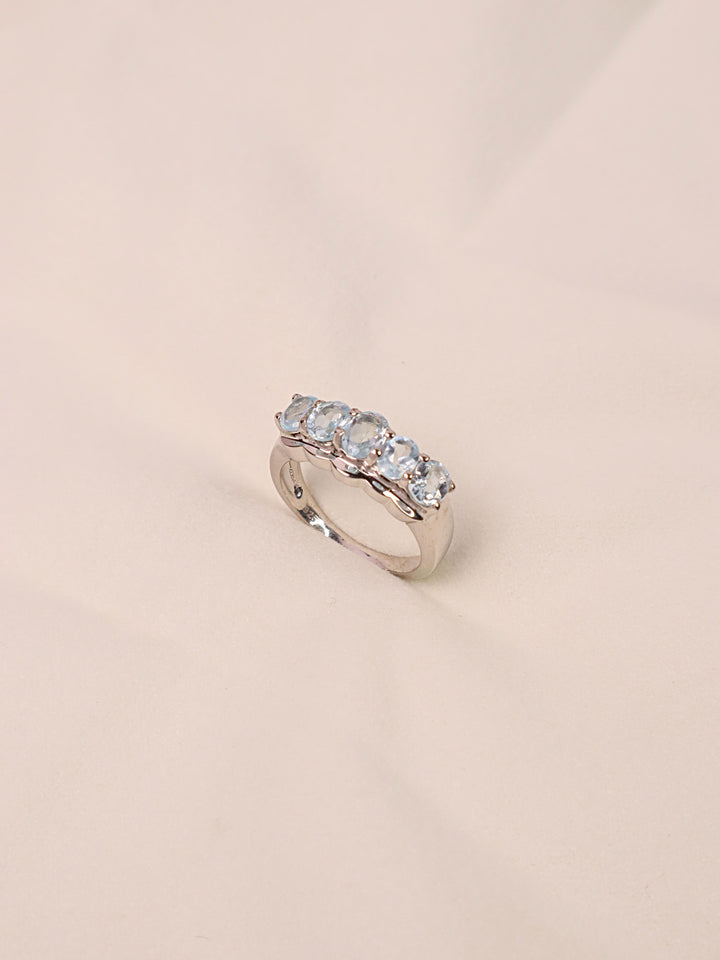 A sterling silver ring with a central blue topaz gemstone surrounded by smaller clear crystals, set on a thin silver band.