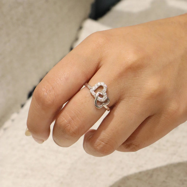 Hand wearing a silver heart-shaped ring on a neutral background