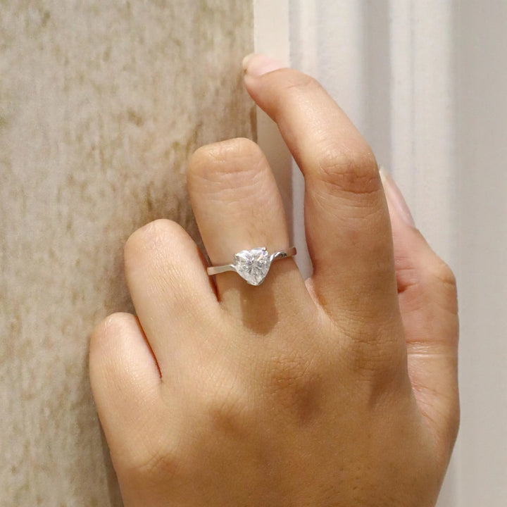 Hand wearing a silver ring with a heart-shaped diamond on a neutral background